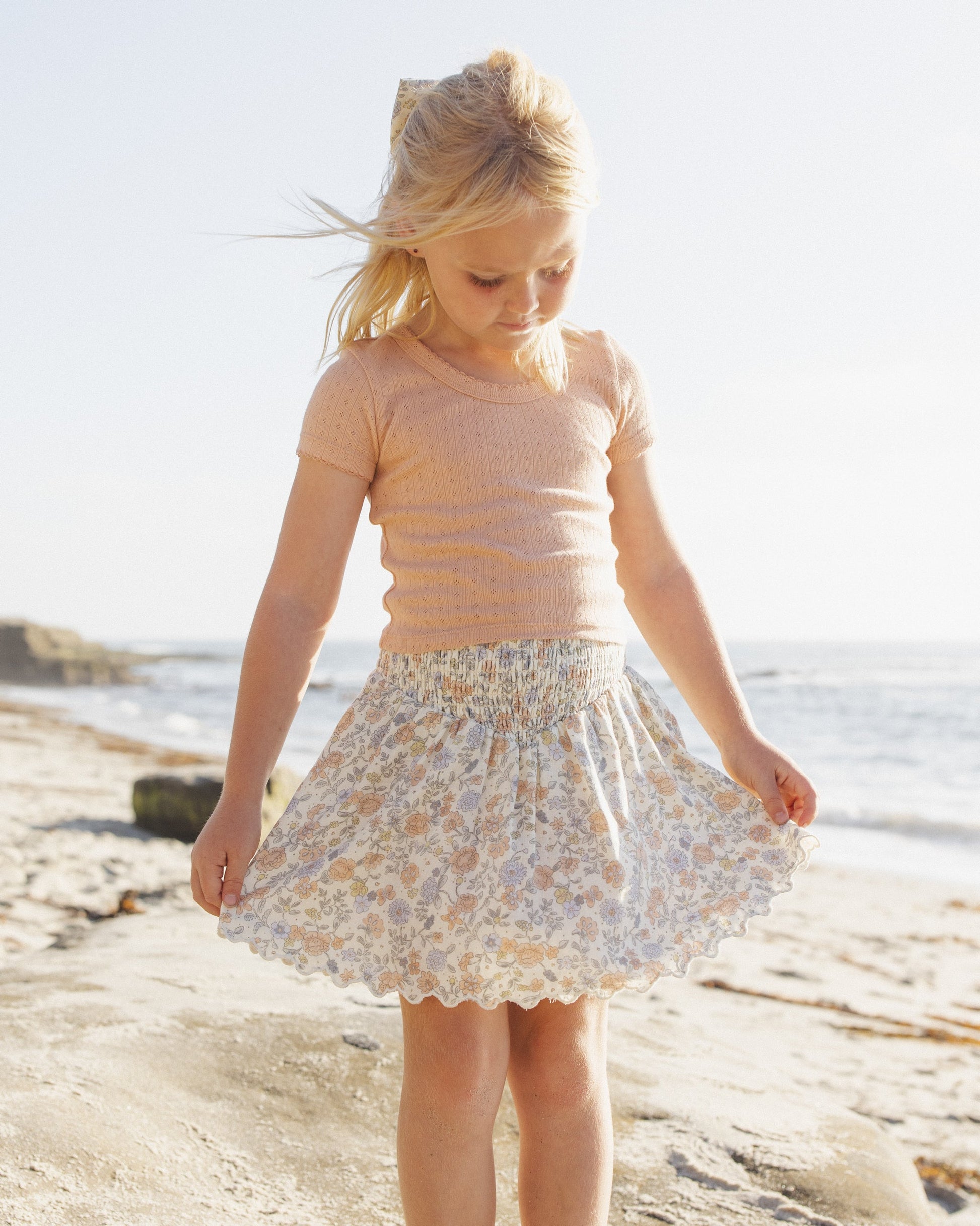 A young girl stands on a sandy beach, looking down and holding the edges of her Rylee & Cru Girls' Smocked V Mini Skirt, with the ocean and sky in the background.