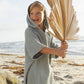 A young child in a sea green Rylee & Cru Kids' Beach Poncho stands smiling on the sand, holding a large dried palm leaf with the ocean in the background.