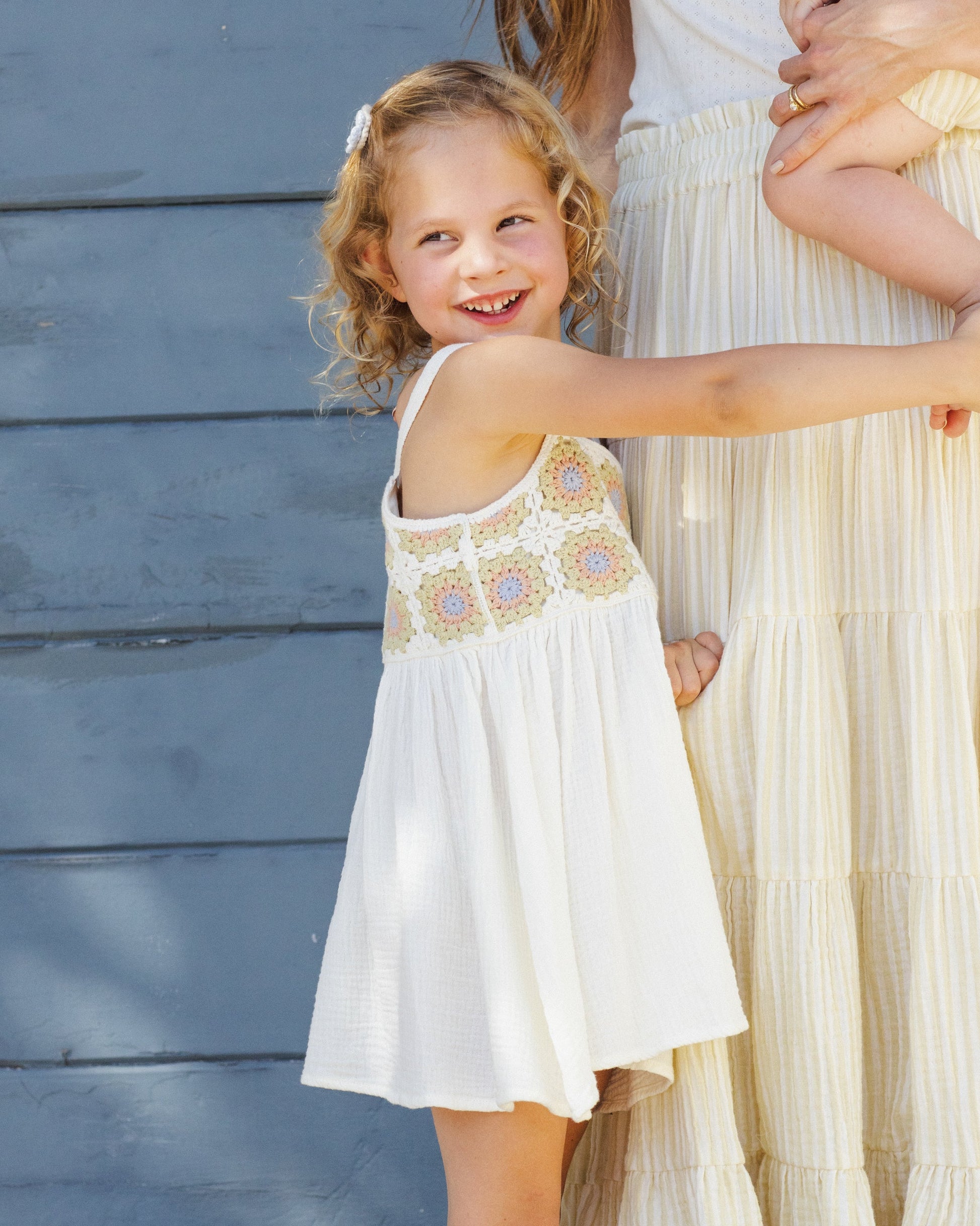 A young girl wearing the Rylee & Cru Girls' Crochet Sundress smiles and points beside an adult in cream, both showcasing vintage charm against a blue wall.