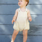 A toddler with short light brown hair wears a Rylee & Cru Baby Lou Romper in citron stripe, standing barefoot on concrete in front of a blue wooden wall.