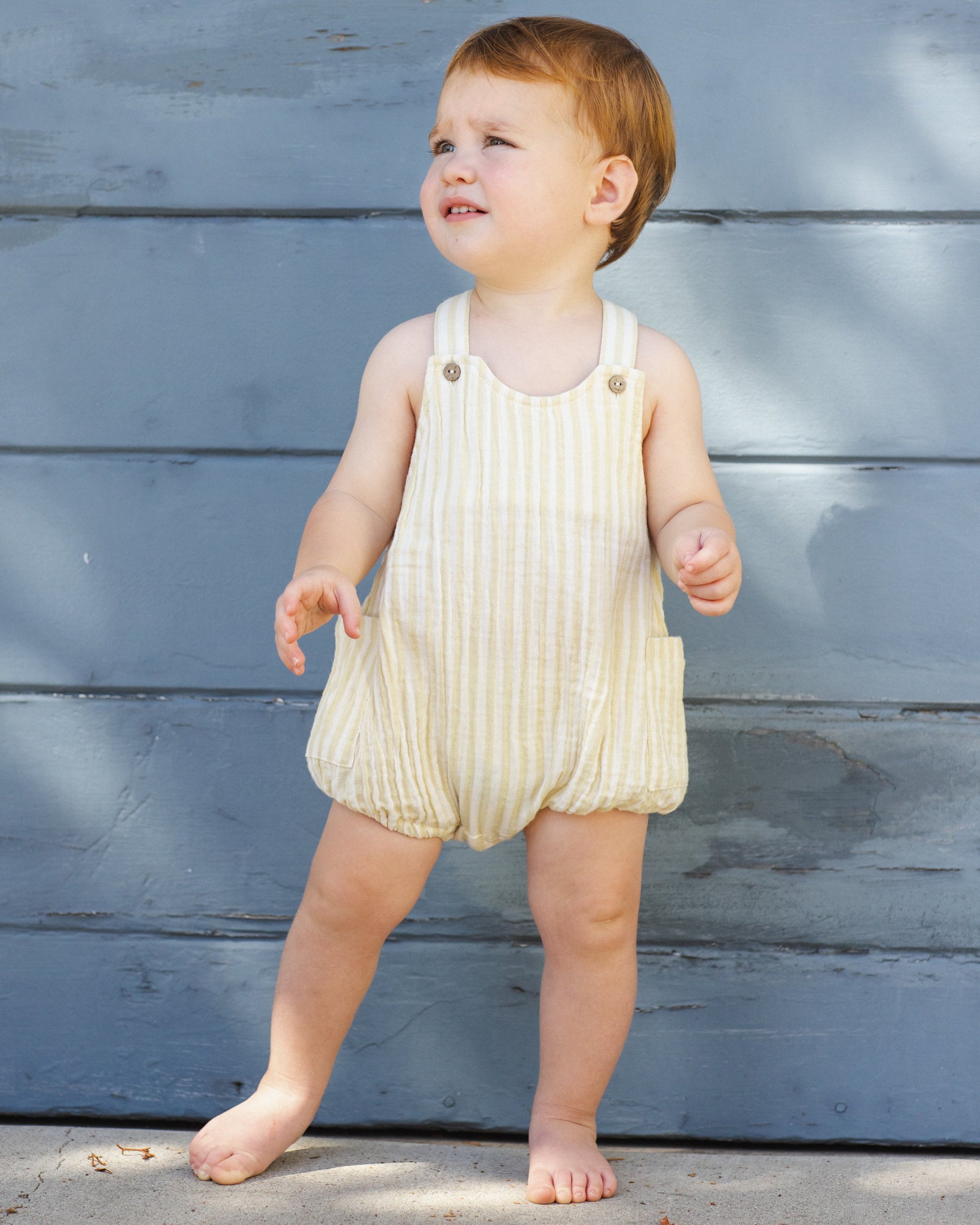 A toddler with short light brown hair wears a Rylee & Cru Baby Lou Romper in citron stripe, standing barefoot on concrete in front of a blue wooden wall.
