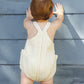 A young child wearing a Rylee & Cru Baby Lou Romper in citron stripe stands facing and touching a blue wooden wall.