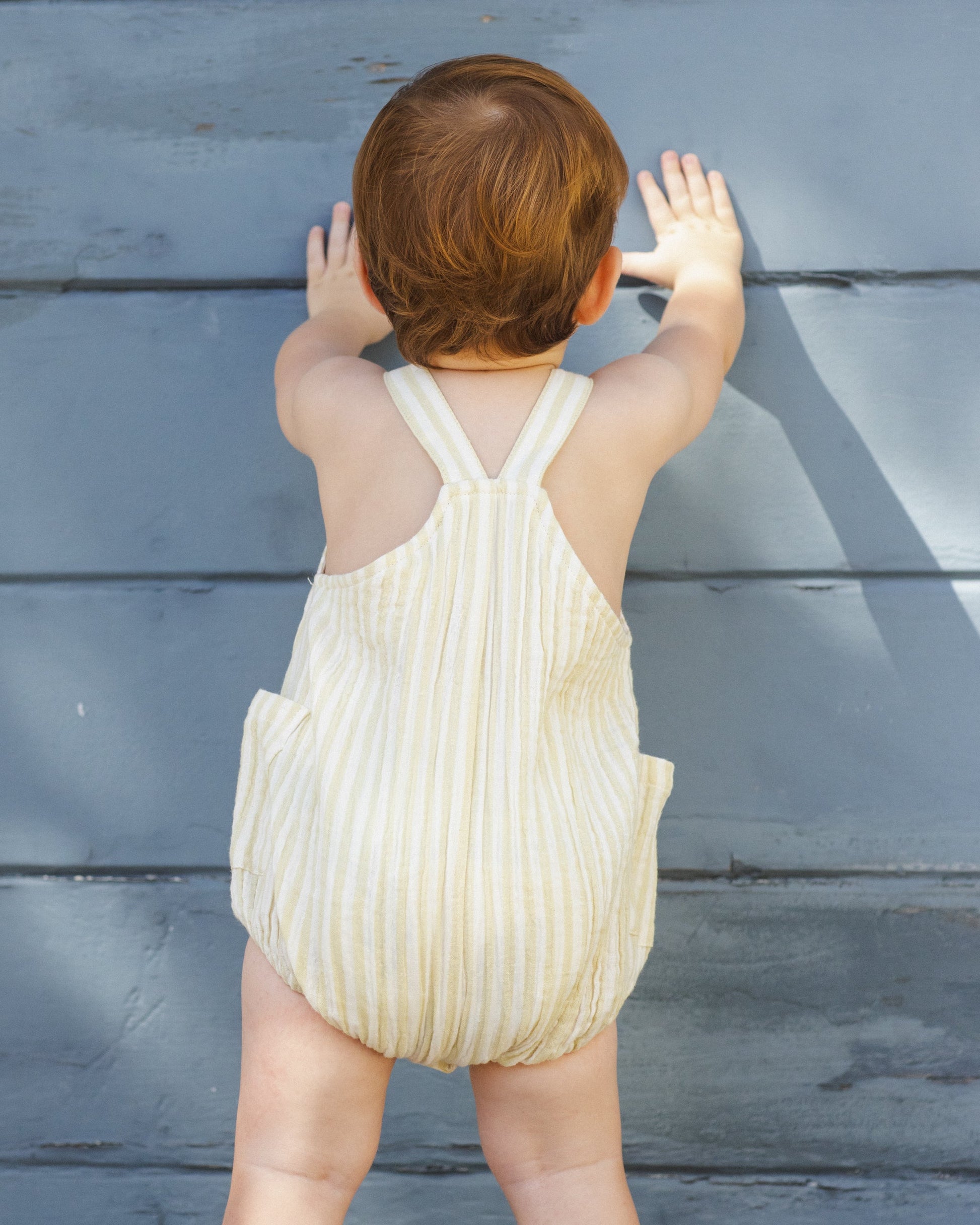 A young child wearing a Rylee & Cru Baby Lou Romper in citron stripe stands facing and touching a blue wooden wall.