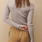 A person wearing the TWP Knit Wool Crewneck, a luxurious grey long-sleeve top by TWP, and beige pants, with their back to the camera and one hand behind their back.