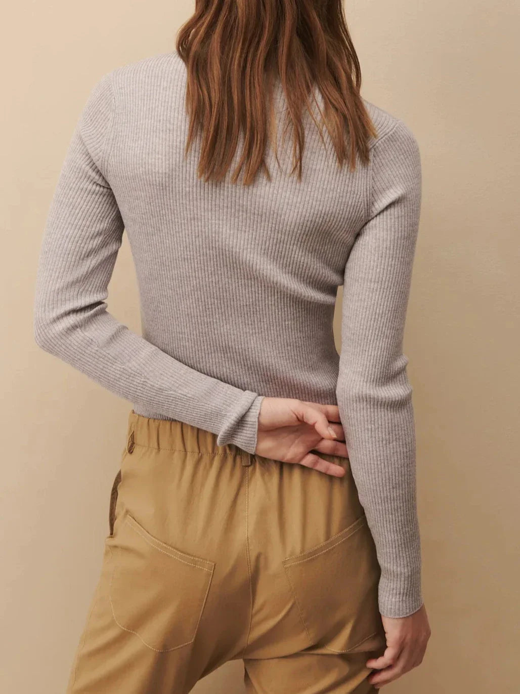 A person wearing the TWP Knit Wool Crewneck, a luxurious grey long-sleeve top by TWP, and beige pants, with their back to the camera and one hand behind their back.