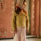 A young girl in a headscarf and knitted sweater stands indoors against a pink, floral-painted wall, looking to the side, dressed in Louise Misha’s Girls’ Salia Pants—cozy ribbed knit pants that add comfort and style to her outfit.