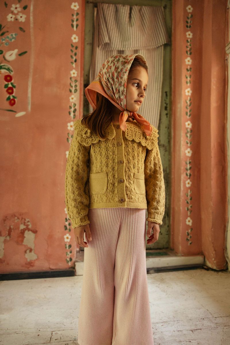 A young girl in a headscarf and knitted sweater stands indoors against a pink, floral-painted wall, looking to the side, dressed in Louise Misha’s Girls’ Salia Pants—cozy ribbed knit pants that add comfort and style to her outfit.