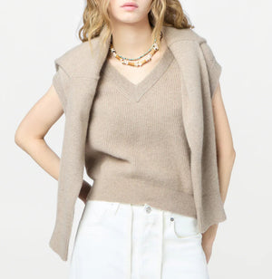 A woman wears the Rib Vest by Guest in Residence in beige, styled with a matching sweater over her shoulders, white pants, and a beaded necklace—an effortlessly chic look.