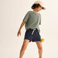 A young child wearing Cozmo Boys' Root Terry Shorts, featuring an elastic waistband and adjustable drawstring, stands with a yellow ball between their knees against a plain background.