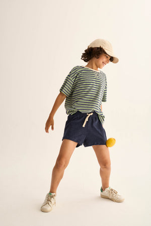 A young child wearing Cozmo Boys' Root Terry Shorts, featuring an elastic waistband and adjustable drawstring, stands with a yellow ball between their knees against a plain background.
