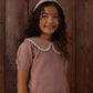 A young girl with long curly hair stands smiling in front of a wooden wall, wearing the Rylee & Cru Girls’ Pointelle Collared Top in pink with a matching headband.