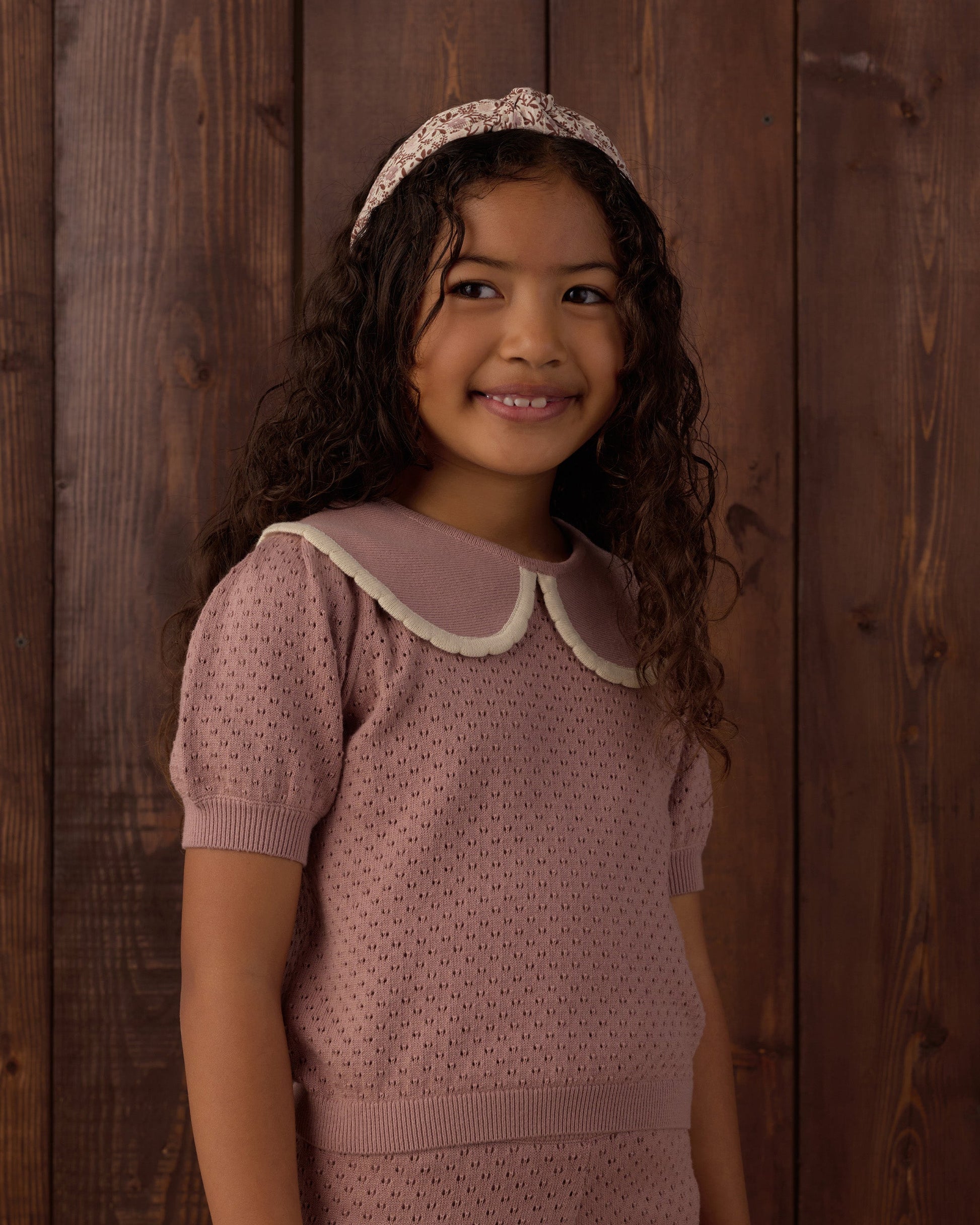 A young girl with long curly hair stands smiling in front of a wooden wall, wearing the Rylee & Cru Girls’ Pointelle Collared Top in pink with a matching headband.