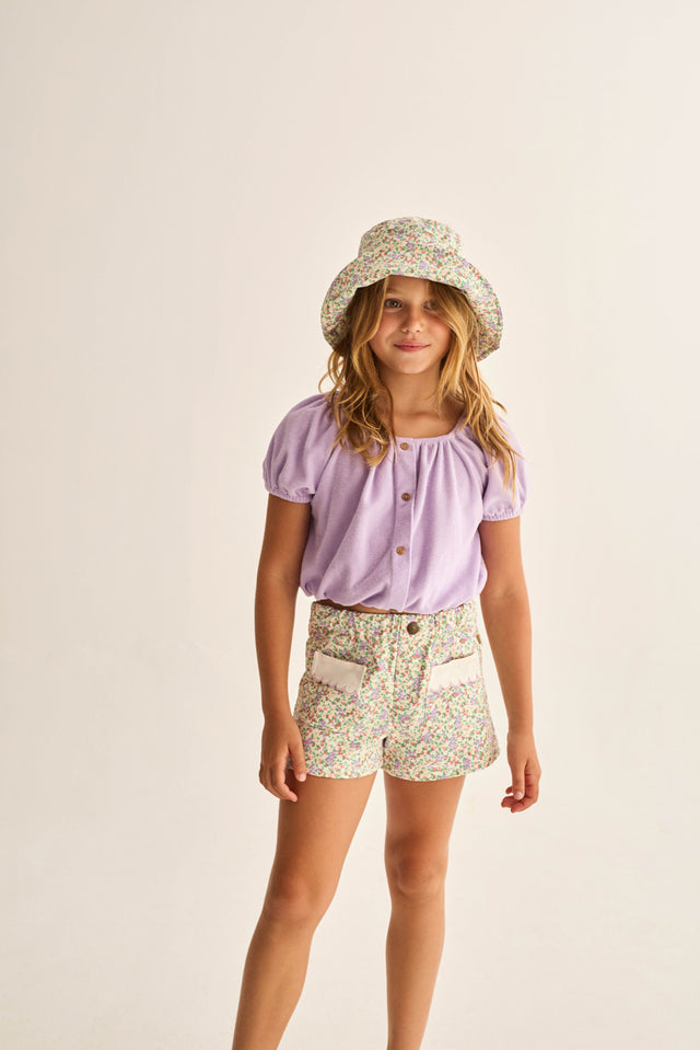 A young girl stands against a plain background, wearing the Cozmo Girls' Judy Terry Top in a cropped silhouette with a boat neckline, paired with a floral bucket hat and matching floral shorts featuring pockets.