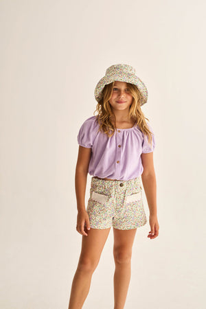 A young girl stands against a plain background, wearing the Cozmo Girls' Judy Terry Top in a cropped silhouette with a boat neckline, paired with a floral bucket hat and matching floral shorts featuring pockets.