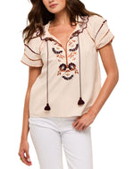 A woman wears the Figue Rosie Top, a light short-sleeve blouse featuring floral embroidery, a smocked neckline, tassel ties, and paired with white pants.