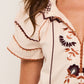 A close-up of a person wearing the Figue Rosie Top, a cream blouse with brown, orange, and maroon floral embroidery, smocked neckline, and decorative sleeve stitching.