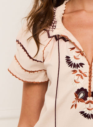 A close-up of a person wearing the Figue Rosie Top, a cream blouse with brown, orange, and maroon floral embroidery, smocked neckline, and decorative sleeve stitching.