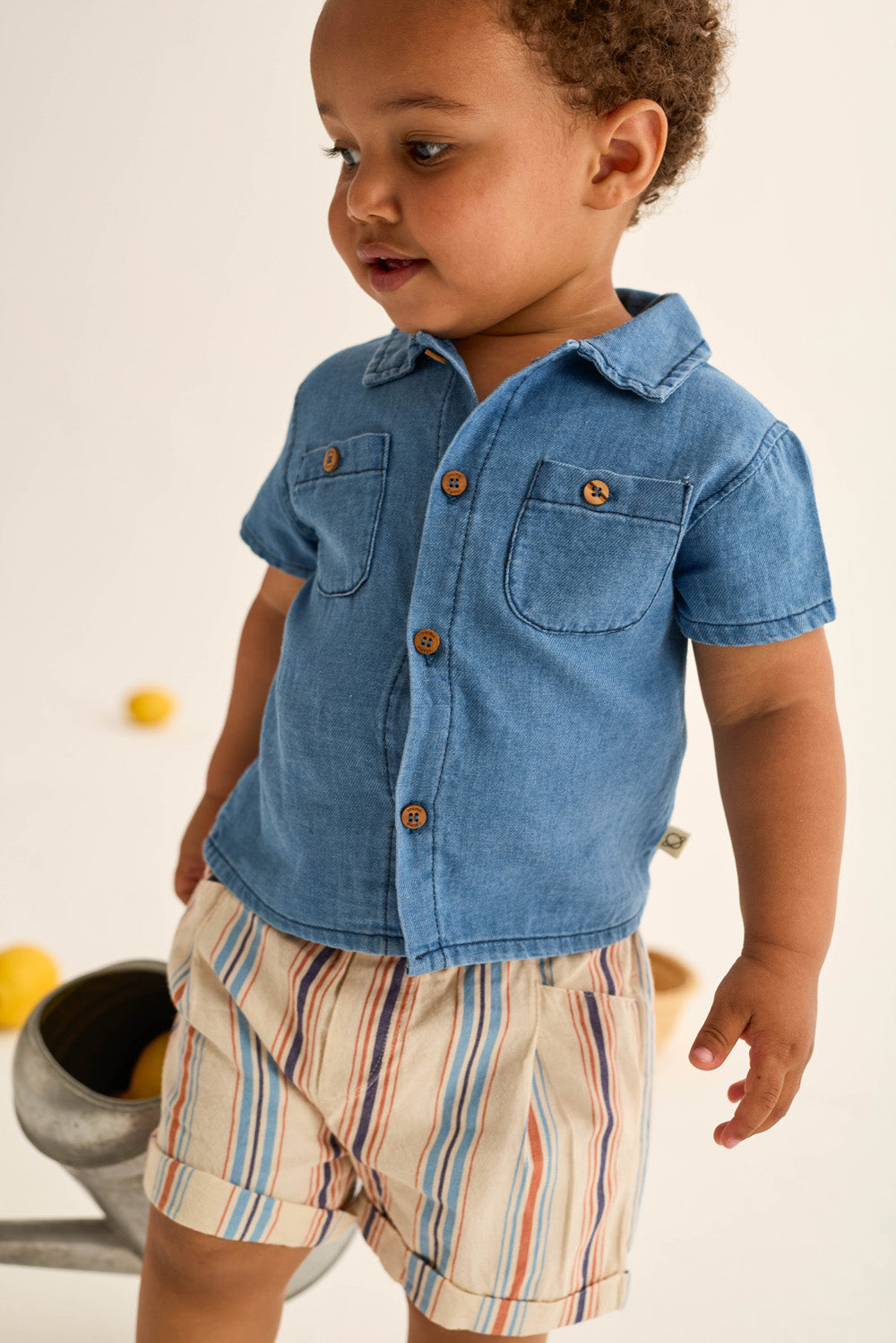 A toddler boy wears Cozmo's Baby Boys' Roy Striped Shorts and a short-sleeve cotton denim shirt with wooden buttons, standing indoors near a watering can and lemons.