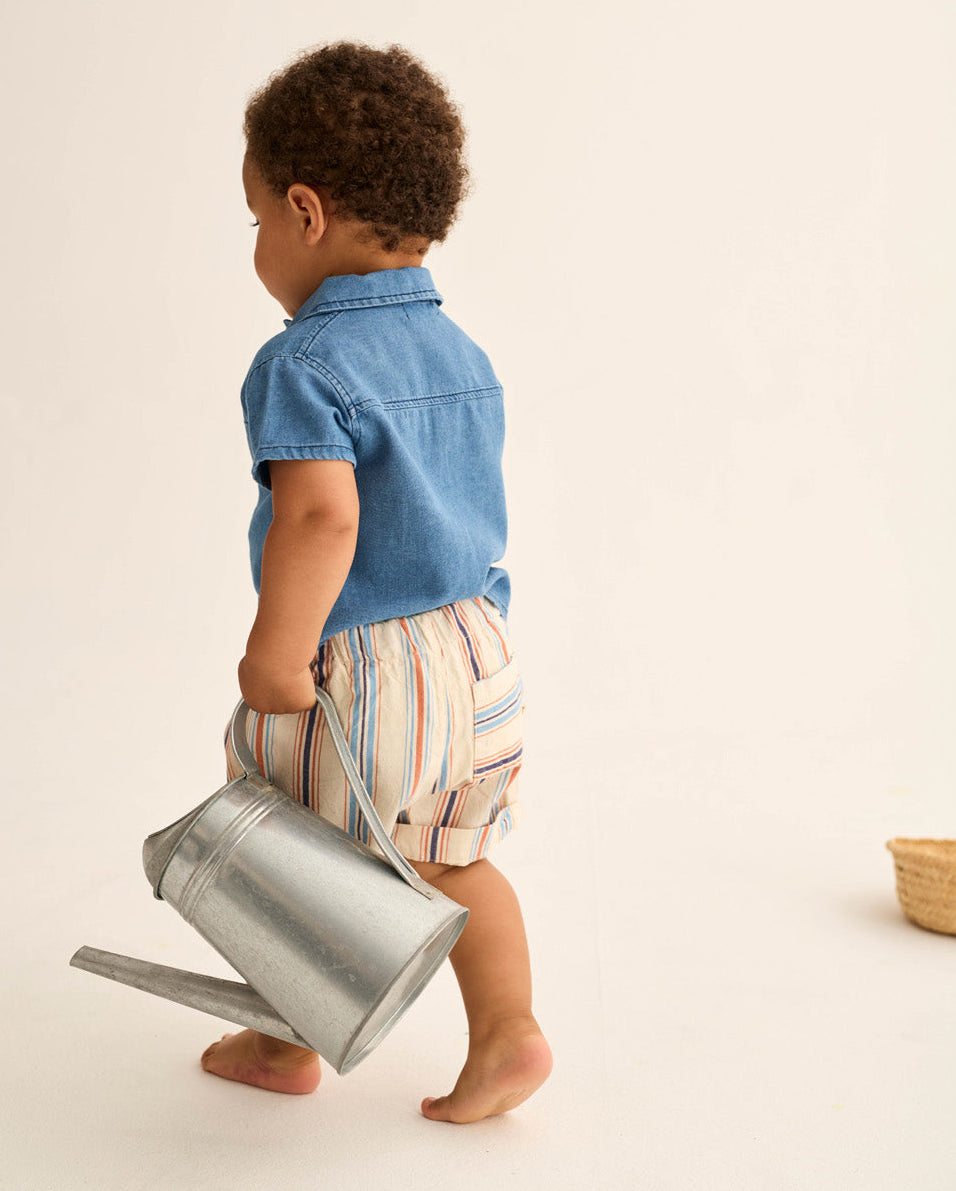 A young toddler boy with short curly hair walks barefoot, wearing a blue shirt and Cozmo Baby Boys' Roy Striped Shorts, while holding a metal watering can.