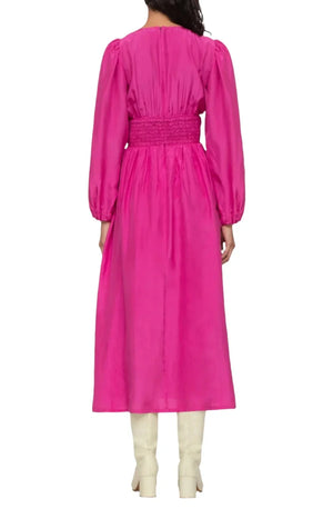 A person stands facing away, wearing the Sea NY Fabiola Dress in a vibrant pink silk habotai fabric, which features a cinched waist and is paired with knee-high white boots.