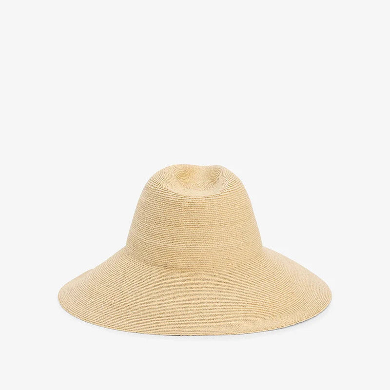 The Janessa Leone Colette Hat is a wide-brim raffia straw hat with a rounded crown, stylishly displayed on a plain white background for chic sun protection.