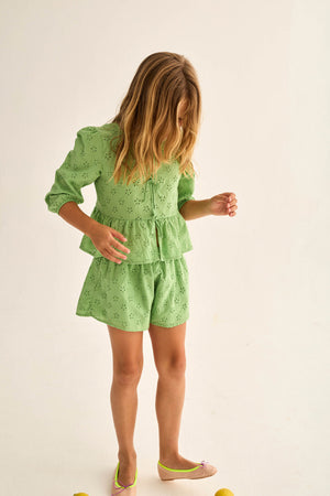 A young girl with long hair wears Cozmo's Girls' Ruby Eyelet Shorts, featuring an elastic waistband and Swiss embroidery, paired with a light green eyelet top, standing barefoot on a plain light background.