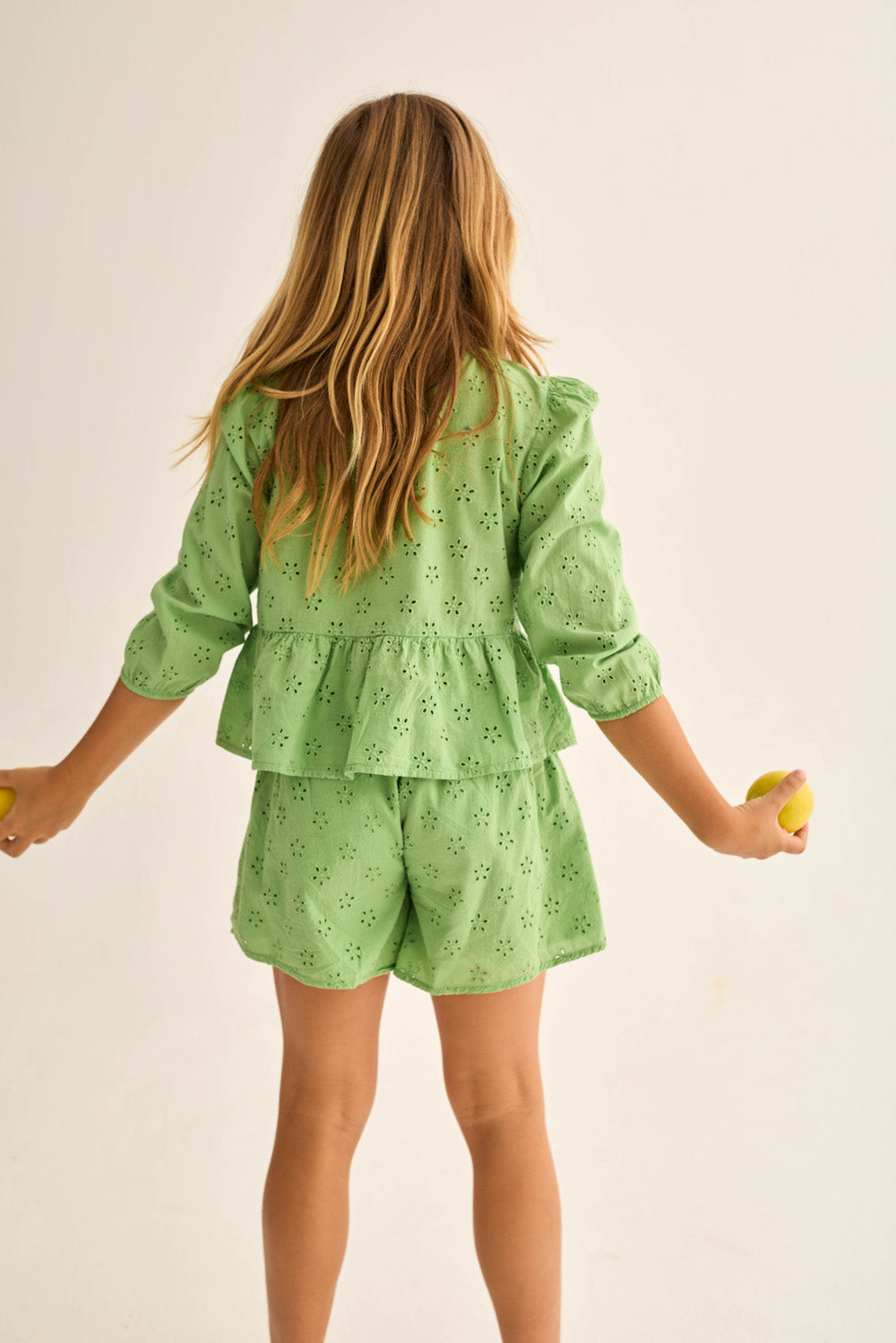 A young girl with long hair, seen from behind, wears Cozmo's Girls' Ruby Eyelet Shorts and a green top while holding two yellow balls against a plain background.