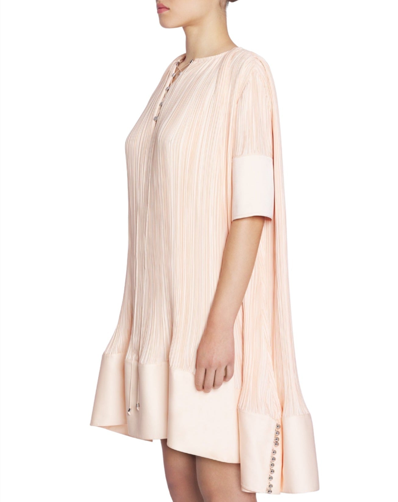 Person wearing a Lanvin blush pink, loose-fitting, knee-length Lanvin Short Sleeve Cascade Dress with long sleeves and a high collar, standing sideways. The pleated dress showcases a gentle cascade effect that adds an elegant touch to the overall look.