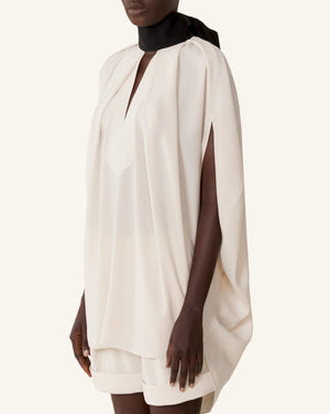 A person wears the Lanvin Knotted High Neck Cape—a loose, off-white cape-style top with slit sleeves and a black grosgrain bow at the neck—paired with matching shorts for a chic Haute Couture look.