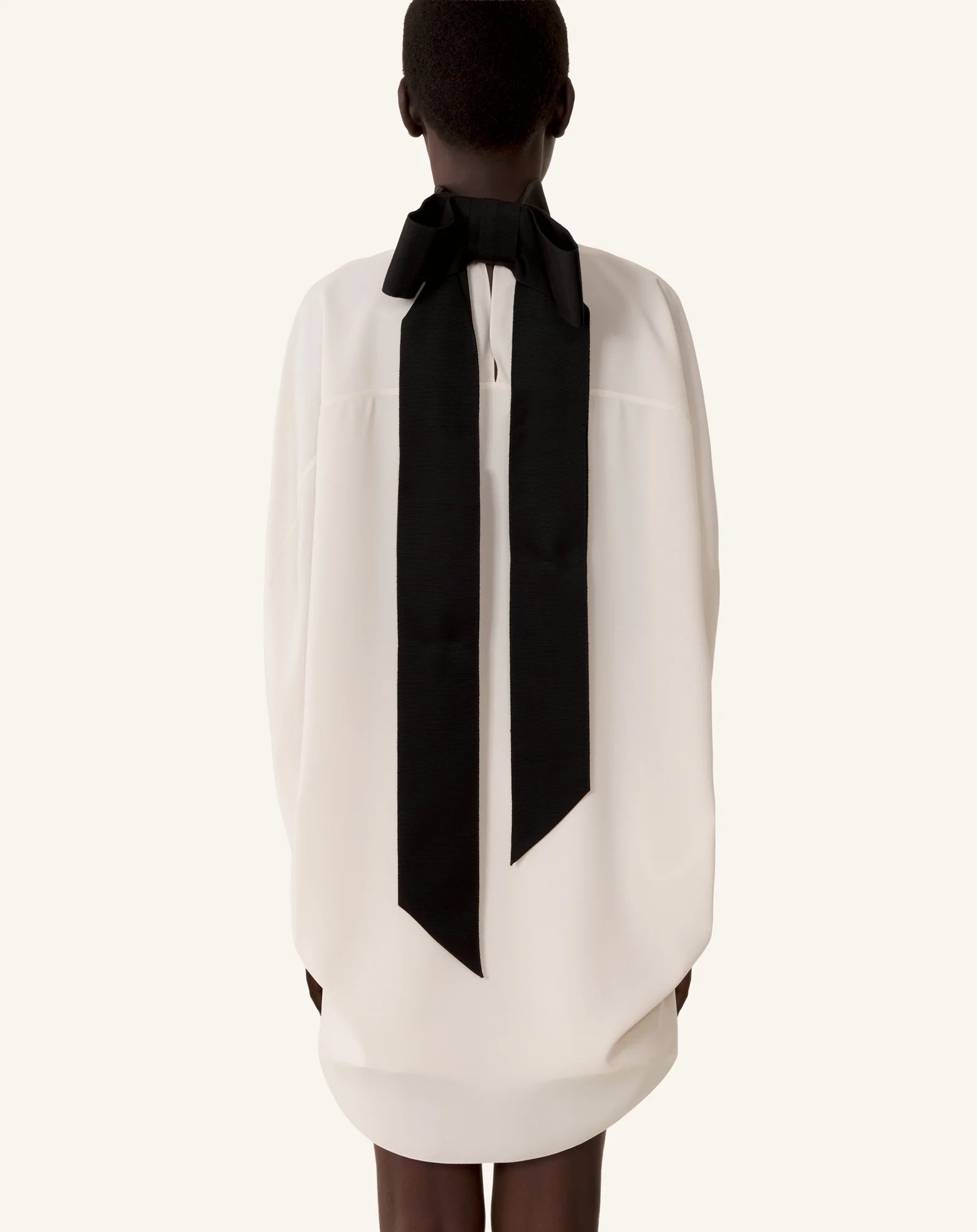 A person stands with their back to the camera, wearing Lanvin’s Knotted High Neck Cape in white, featuring a dramatic grosgrain bow at the neck.