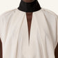 A person wearing Lanvin’s Knotted High Neck Cape, featuring a high black collar, white loose fit, deep V pleat at the front, and grosgrain bow for a Haute Couture finish.