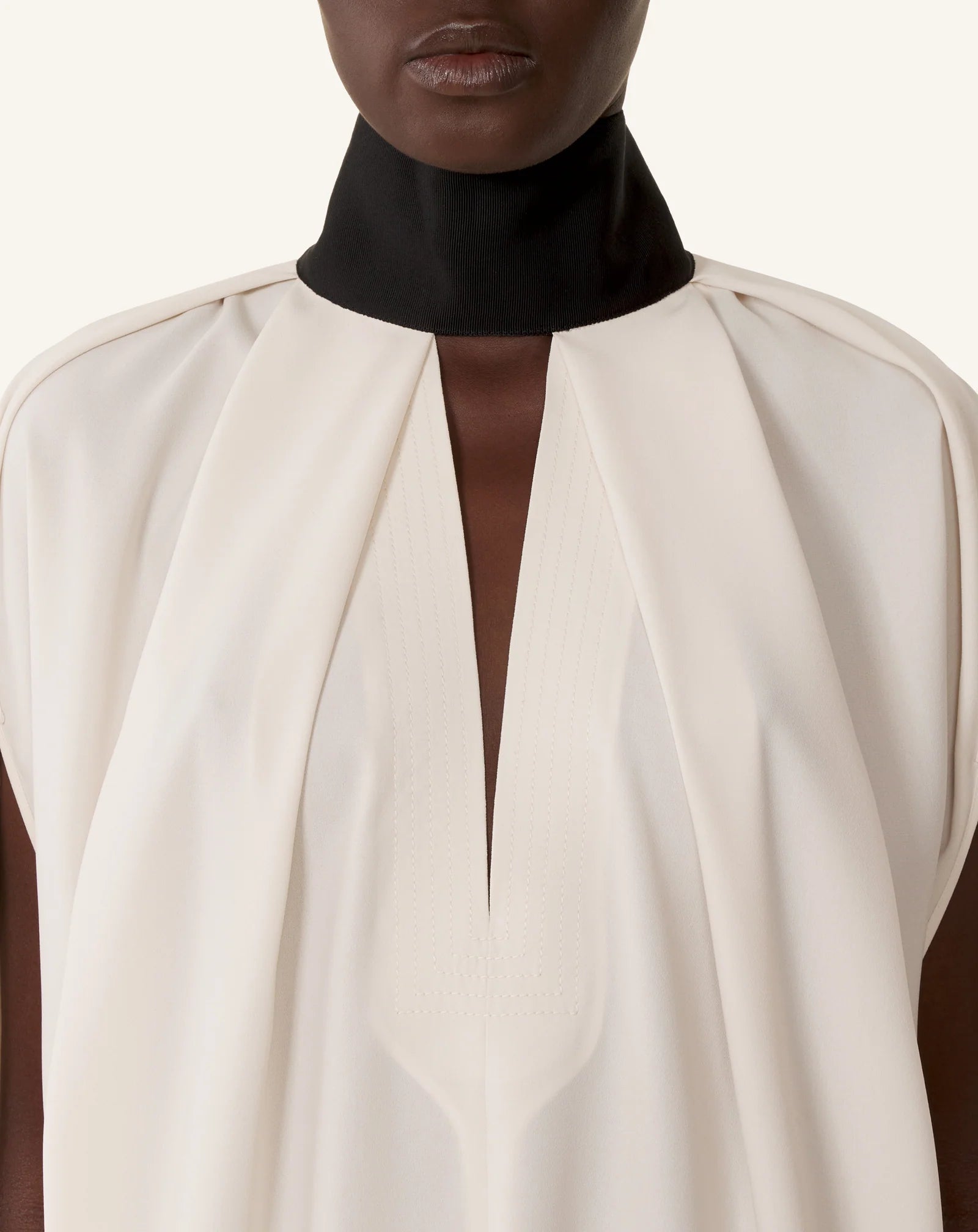A person wearing Lanvin’s Knotted High Neck Cape, featuring a high black collar, white loose fit, deep V pleat at the front, and grosgrain bow for a Haute Couture finish.