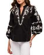 A woman wears the Figue Rylie Top, a black blouse with white floral embroidery, blouson sleeves, and a band collar, styled with white pants.