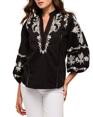 A woman wears the Figue Rylie Top, a black blouse with white floral embroidery, blouson sleeves, and a band collar, styled with white pants.
