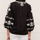 A woman is shown from the back wearing the Figue Rylie Top, a black blouse with white floral embroidery, blouson sleeves, and a relaxed fit, paired with white pants.