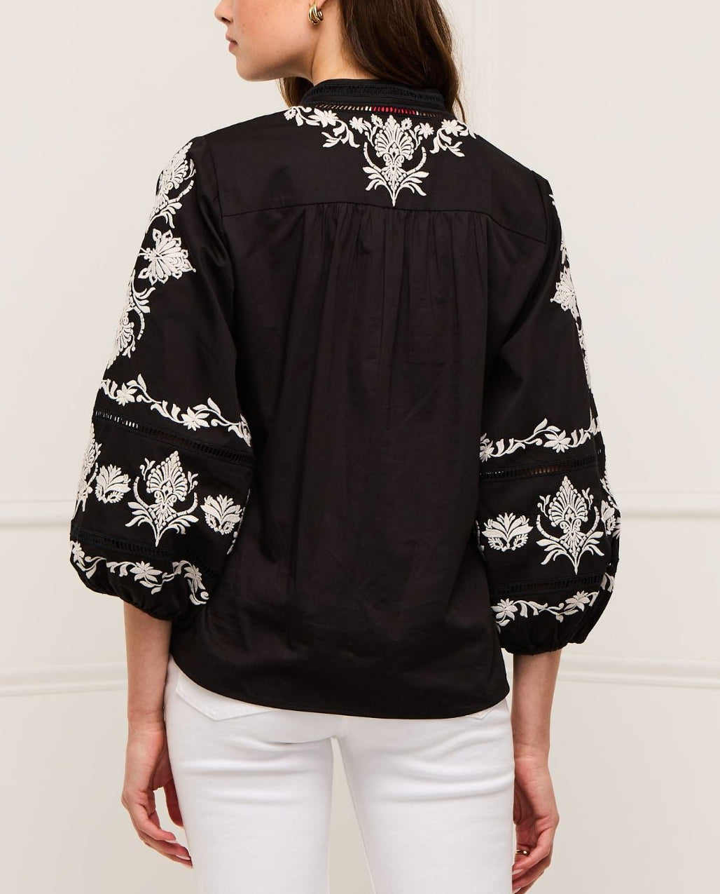A woman is shown from the back wearing the Figue Rylie Top, a black blouse with white floral embroidery, blouson sleeves, and a relaxed fit, paired with white pants.
