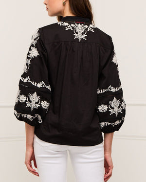 A woman is shown from the back wearing the Figue Rylie Top, a black blouse with white floral embroidery, blouson sleeves, and a relaxed fit, paired with white pants.