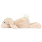 A large, plush Jellycat Smudge Rabbit in beige, with oatmeal fur and a white tail, is lying on its stomach and facing left.