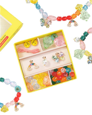 The Super Smalls Make It Rainbow Mini Bead Kit includes vibrant beads, smiley face accents, rainbow charms, elastic string, earrings, and crystals—perfectly arranged in a yellow box for creative jewelry-making fun.