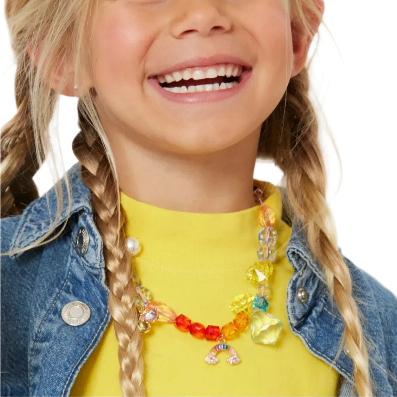 A smiling child with blonde braids wears a yellow shirt, denim jacket, and a colorful chunky necklace made with vibrant beads from the Super Smalls Make It Rainbow Mini Bead Kit.