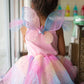 A child sits by the window, dressed in the Great Pretenders Rainbow Fairy Dress & Wings, with shimmering organza catching the light beautifully.