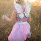 A child with curly blonde hair is gleefully standing in the field, wearing the Great Pretenders Rainbow Sequins Skirt, Wings & Wand set from Great Pretenders.