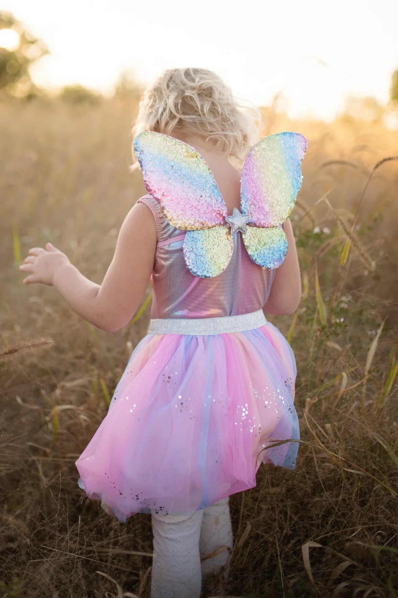 A child with curly blonde hair is gleefully standing in the field, wearing the Great Pretenders Rainbow Sequins Skirt, Wings & Wand set from Great Pretenders.