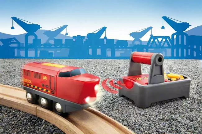 A BRIO World Remote Control Engine toy train by Brio glides smoothly on wooden tracks, set against a backdrop of crane and industrial structure silhouettes.