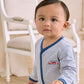 A toddler stands indoors in front of a white upholstered chair, looking slightly to the side, wearing the Kissy Kissy Baby Boys’ Rescue Raid Striped Footie.