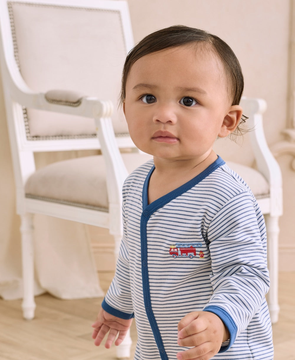 A toddler stands indoors in front of a white upholstered chair, looking slightly to the side, wearing the Kissy Kissy Baby Boys’ Rescue Raid Striped Footie.