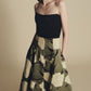 A woman stands against a plain beige background wearing the Lela Rose Parker Floral Velvet Skirt, featuring a green hue with large white floral patterns and a strapless black top.