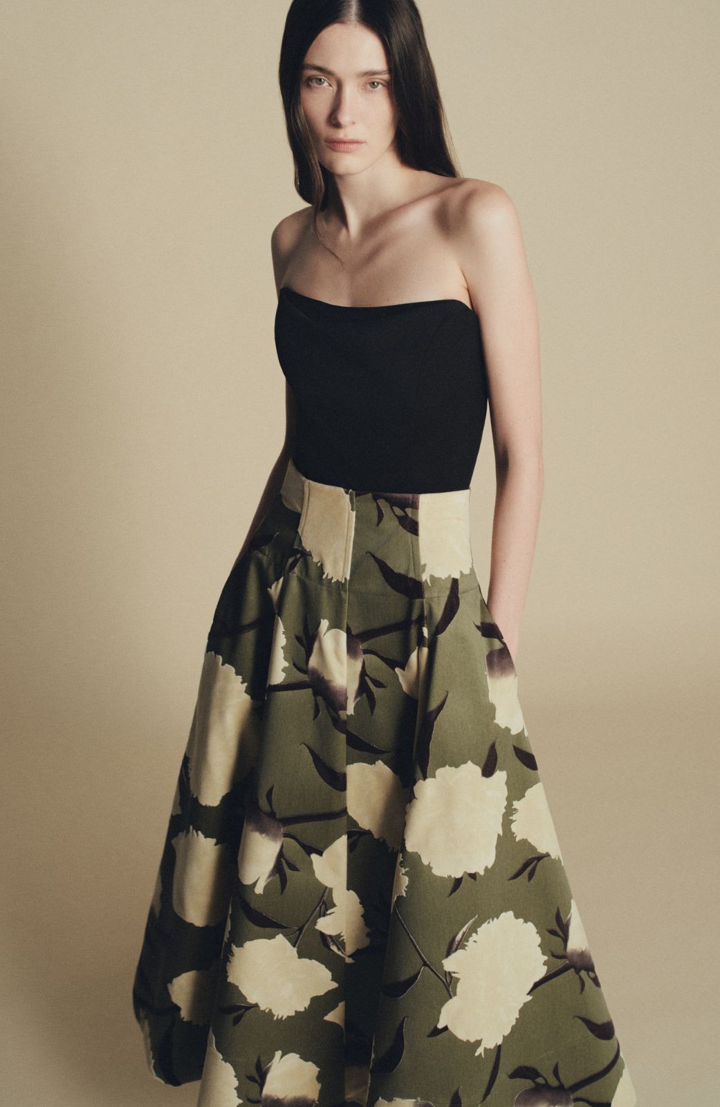 A woman stands against a plain beige background wearing the Lela Rose Parker Floral Velvet Skirt, featuring a green hue with large white floral patterns and a strapless black top.
