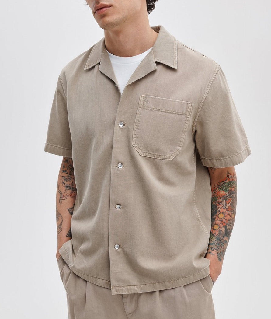 A person in a Rag & Bone Resort Denim Shirt over a white tee, paired with beige pants and showcasing arm tattoos, exemplifies effortless resort style.