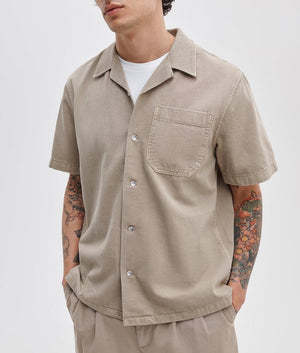 A person in a Rag & Bone Resort Denim Shirt over a white tee, paired with beige pants and showcasing arm tattoos, exemplifies effortless resort style.